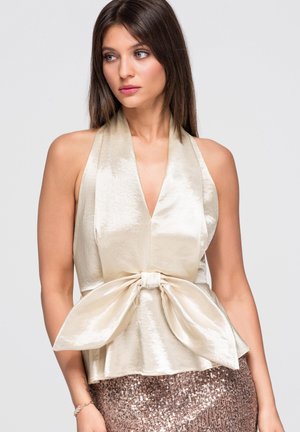 Donna con capelli castani lisci indossa un top crema senza maniche con un grande fiocco e una gonna scintillante con paillettes, guarda di lato.