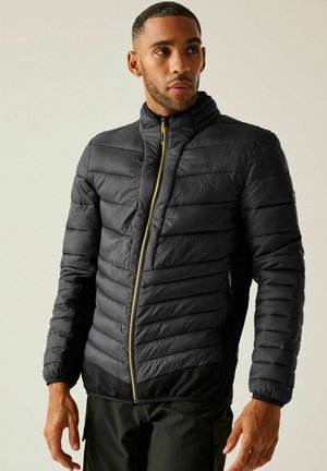 Hombre con chaqueta acolchada negra con cremallera amarilla, de pie con los brazos relajados, mirando ligeramente hacia la derecha sobre un fondo liso.