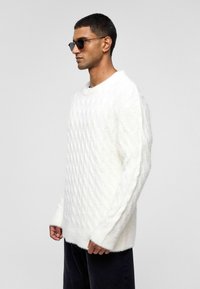 Weißer Kabelstrick-Pullover mit Rundhalsausschnitt und langen Ärmeln, mit gerippten Bündchen und leichter Passform. Getragen mit einer schwarzen Sonnenbrille.
