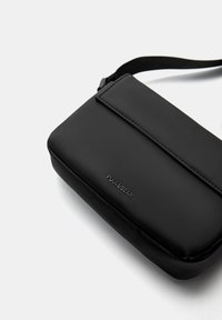 Sort syntetisk crossbody taske med en glat tekstur, rektangulær form, klaplukning og præget logo på forsiden. Justerbar rem inkluderet.