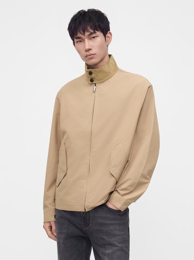 Jeune homme portant une veste beige zippée avec un col haut et un jean noir, debout avec une main dans la poche sur un fond uni.