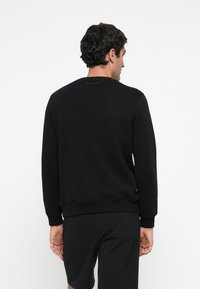 Uomo che indossa una felpa nera a maniche lunghe e pantaloni corti neri, in piedi con le spalle rivolte verso l'osservatore su uno sfondo bianco.