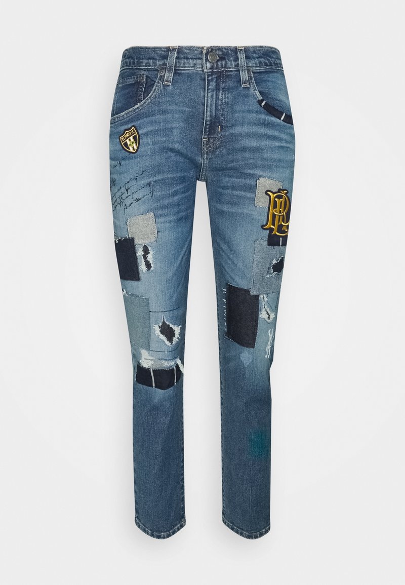Jean en denim bleu délavé avec une coupe slim, présentant divers patchs, des détails usés et des logos brodés sur le devant.