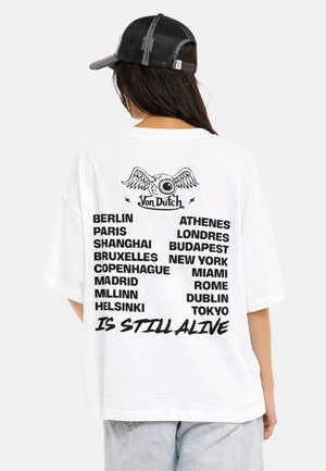 Femme portant un t-shirt blanc Von Dutch listant des villes mondiales et le slogan "EST TOUJOURS VIVANT" dans le dos, assorti d'une casquette noire en mesh et d'un jean clair.