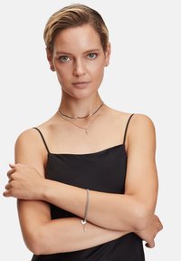 Gioielli in argento con una collana a strati in design curvo e a catena, abbinati a un bracciale, indossati su un braccio incrociato su un vestito nero.