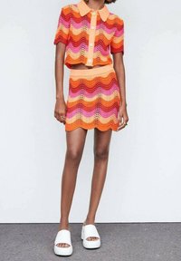 Femme portant un ensemble en crochet deux pièces aux motifs ondulés colorés orange, rose et rouge, avec un haut à col et des manches courtes, ainsi qu'une mini-jupe, et des sandales à plateforme blanches.