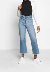 Hochtaillierte Jeans aus Denim in Hellblau mit einer cropped, ausgestellten Silhouette und fransigem Saum, kombiniert mit einem schwarzen Langarmshirt und weißen Sneakern.