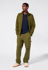 Grüne Jacke mit Reißverschluss über einem marineblauen T-Shirt, kombiniert mit olivgrünen Hosen und beigen Schuhen. Legeres, strukturiertes Design mit Fronttaschen.