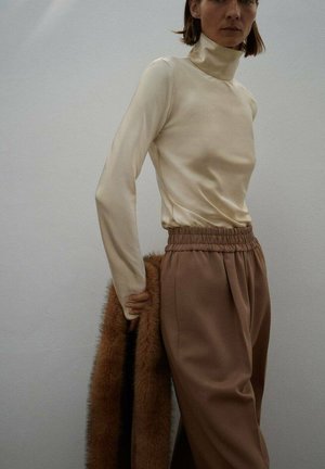 Un haut à col roulé crème à manches longues associé à un pantalon élastique taille haute marron clair, avec un manteau en fausse fourrure marron drapé sur le bras.