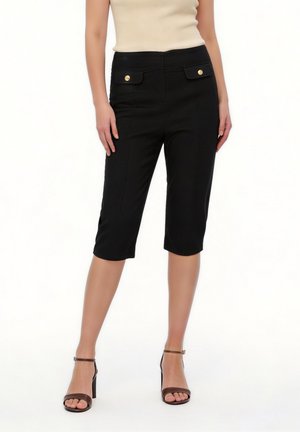 Kobieta ubrana w czarne, dopasowane spodenki do kolan z przednimi kieszeniami na zakładkę i złotymi guzikami, zestawione z beżowym topem bez rękawów i czarnymi szpilkami.