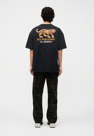 Férfi fekete, túlméretezett pólóban, narancssárga leopárdmintával és „Les Benjamins El Gringo” felirattal a hátán, virágmintás nadrágban és platform cipőben.