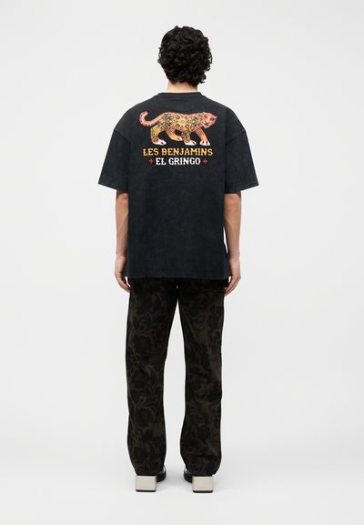 Άνδρας που φοράει μαύρο υπερμεγέθες T-shirt με πορτοκαλί λεοπάρ σχέδιο και κείμενο «Les Benjamins El Gringo» στην πλάτη, παντελόνι με λουλουδάτο μοτίβο και παπούτσια με πλατφόρμα.