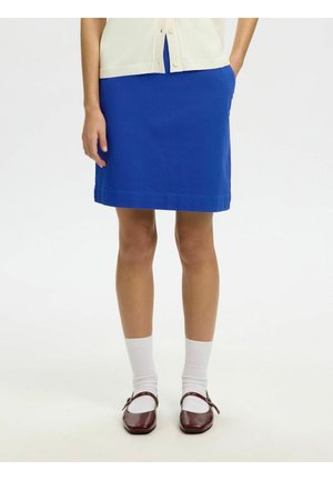 Personne portant une jupe bleue mi-genoux, des chaussettes blanches et des chaussures Mary Jane rouge foncé, debout avec une main dans la poche sur fond blanc.