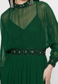 Robe vert émeraude avec des manches longues transparentes, un col haut et une taille ainsi que des poignets ornés de sequins. Présente une jupe plissée pour ajouter de la texture.