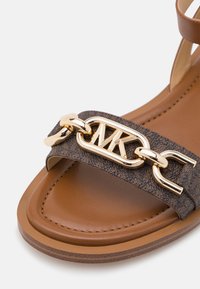Sandale marron avec bride texturée et détail chaîne dorée présentant des lettres entrelacées M et K sur le dessus à l'avant.