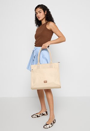 Borsa tote beige con doppi manici, realizzata in materiale liscio. Distintivo patch del logo marrone sul davanti. Abbinata a una gonna blu plissettata.