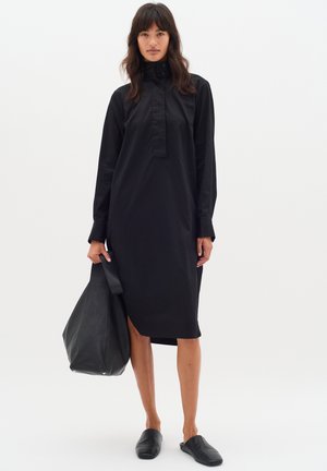 Robe chemise en coton noir avec un col haut, des manches longues et des fentes sur les côtés ; assortie à un sac à main en cuir noir et des chaussures à enfiler.