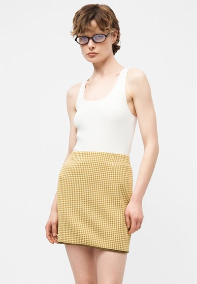 Άτομο με κοντά καστανά μαλλιά και γυαλιά που φοράει λευκό αμάνικο τοπ και κίτρινη καρό μίνι φούστα σε απλό φόντο.