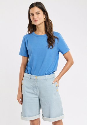 Femme aux longs cheveux foncés portant une chemise bleue à manches courtes et un short rayé bleu clair avec des boutons, debout, une main dans la poche.