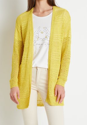 Personne portant un cardigan en maille jaune ouverte par-dessus un t-shirt blanc avec un motif de mains en forme de cœur et un pantalon beige, debout devant un fond clair.