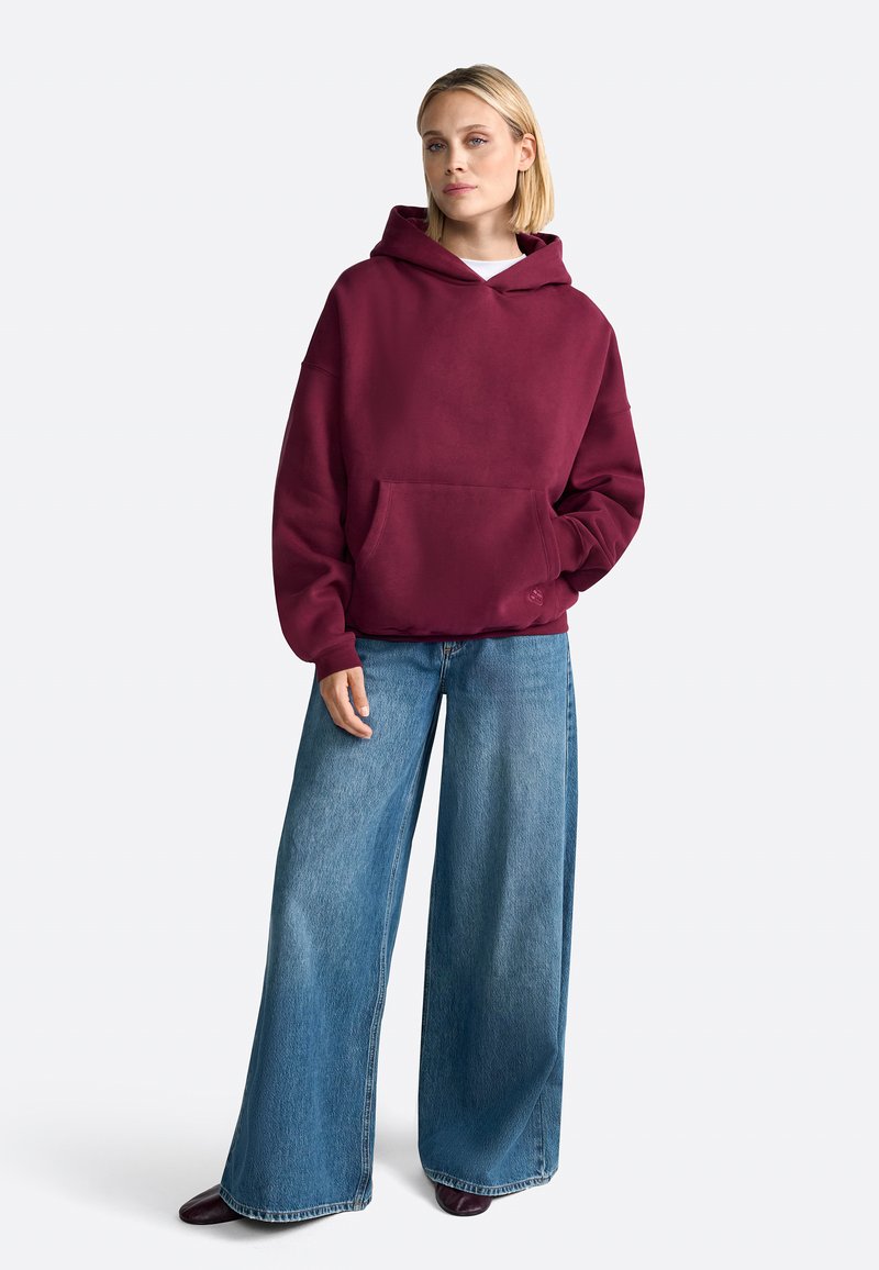 Burgundy hooded sweatshirt with a front pocket, paired with wide-leg blue jeans. Fabric appears soft with a relaxed fit design.