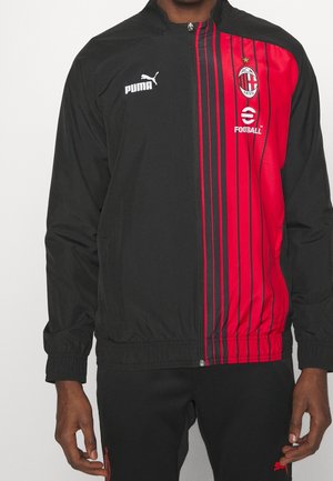 Personne portant une veste de sport zippée noire et rouge de l'AC Milan avec les logos Puma et football, debout devant un fond uni.