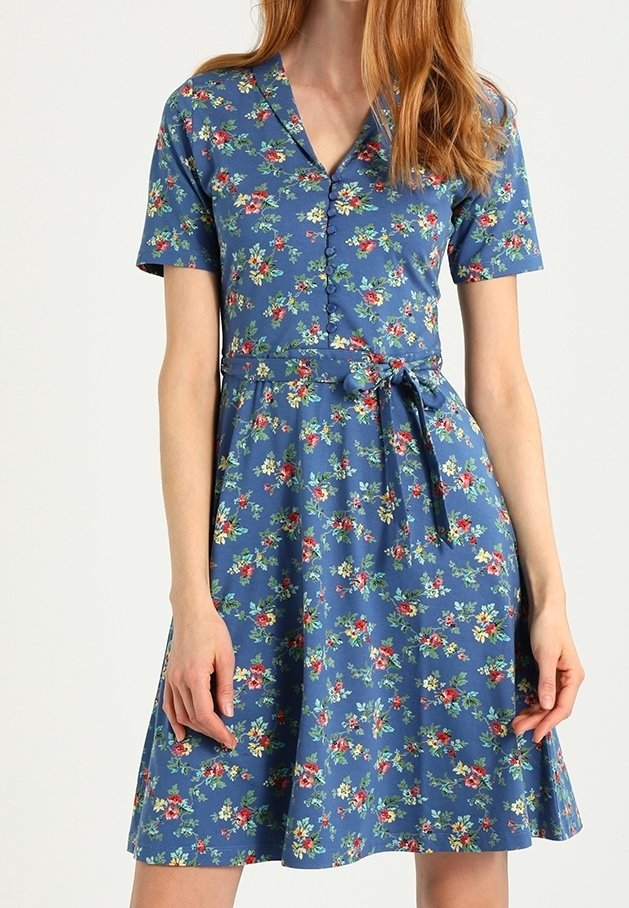 Blå blommig klänning med korta ärmar, knappframsida och ett knytbälte. Har ett litet blommönster med röda och gröna accenter på en blå bakgrund.