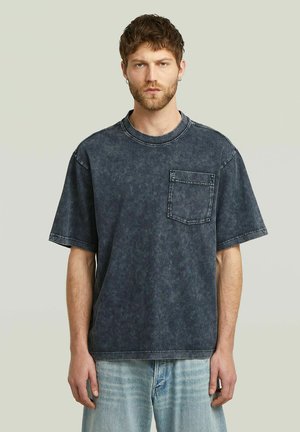 Jonge man met kort haar en baard, gekleed in een vervaagde zwarte oversized T-shirt met een borstzakje en lichtblauwe jeans, staat neutraal.