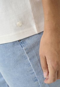 Bílá bavlněná košile s detailním knoflíkem na spodní části, spárovaná se světle modrými džíny. Jasné švy na obou kusech oděvu.