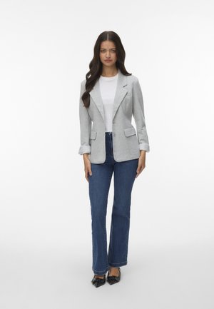 Blazer gris con solapas de pico, dos bolsillos frontales y cierre de botón, combinado con jeans acampanados azules y bailarinas negras de punta.
