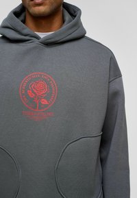 Grijze hoodie met een ronde rode graphic van een roos en tekst, met een zachte textuur, verstelbare capuchon en een ontwerp met een buidelzak aan de voorkant.
