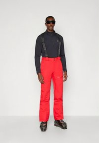 Červené lyžiarske nohavice s nastaviteľnými čiernymi trakmi, vyrobené z vodeodolného materiálu. Nosené s čiernym tričkom s dlhým rukávom a čiernymi lyžiarskymi topánkami.