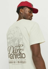 T-shirt in cotone color crema con una grafica dettagliata sul retro che raffigura un sole e delle ali, con la scritta "Dark Manifesto" in caratteri marroni.