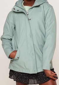 Light green waterproof jacket with a hood, featuring a zip closure, buttoned placket, and side pockets, paired with a black polka dot skirt.