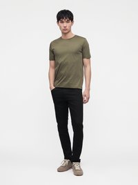 Jonge man staand, draag een olijfgroen T-shirt, zwarte broek en lichtbruine schoenen tegen een effen witte achtergrond.