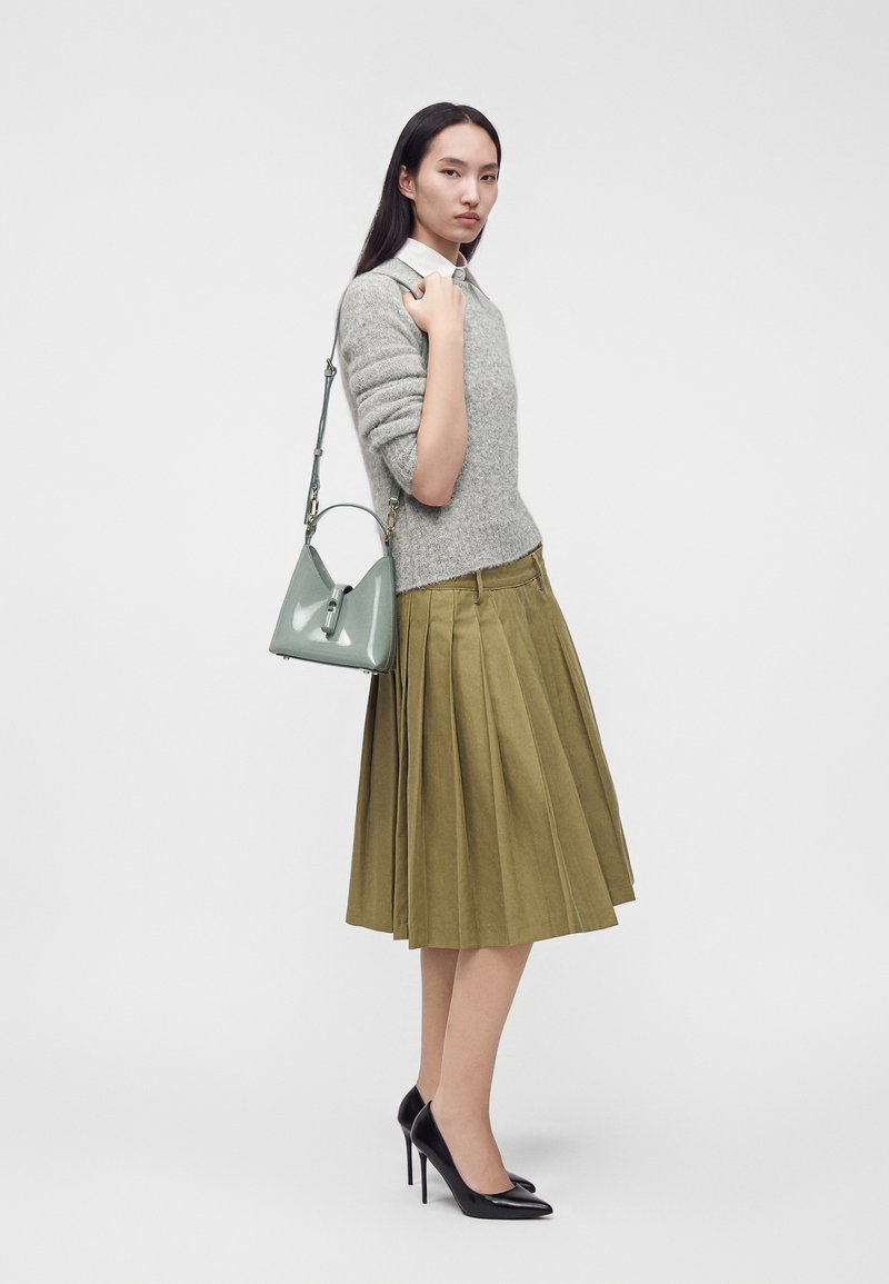 Femme portant un pull gris, une jupe plissée vert olive, des talons hauts noirs, et portant un petit sac à main gris à l'épaule.