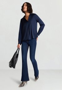 Blouse bleu marine avec un col en V, manches longues et une coupe décontractée, associée à un jean évasé sombre et des talons pointus en cuir façon serpent. Sac à franges noir.