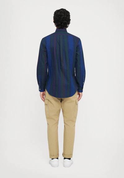 Camisa de manga larga azul oscuro con rayas verticales en verde y rojo, combinada con pantalones cargo caqui, vista desde la parte trasera. Zapatillas blancas.