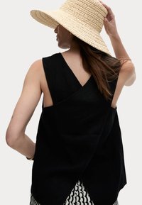 Top negro sin mangas con un diseño de espalda cruzada, que presenta un corte relajado y una tela texturizada. Combinado con un sombrero de paja de ala ancha.