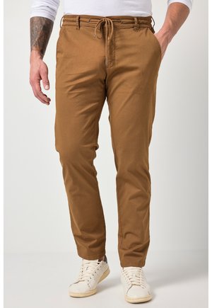 Uomo che indossa pantaloni marroni con coulisse e tasche, e sneakers bianche, in piedi con una mano in tasca contro uno sfondo semplice.