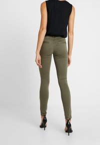 Olive green skinny jeans with a smooth texture and rear pockets, paired with a black sleeveless top and black high-heeled shoes.