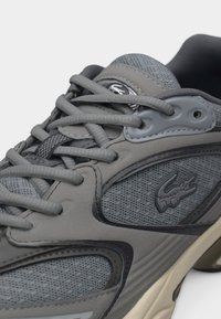 Γκρι αθλητικό παπούτσι με πάνελ από δίχτυ και δέρμα, στρογγυλά γκρι κορδόνια και μαύρο ανάγλυφο λογότυπο κροκόδειλου στο πλάι κοντά στη σόλα.