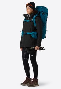 Giacca nera da esterno con spallacci blu, zaino turchese, leggings neri e scarpe da trekking marroni. Tiene bastoncini da trekking, indossa un berretto e occhiali da sole.