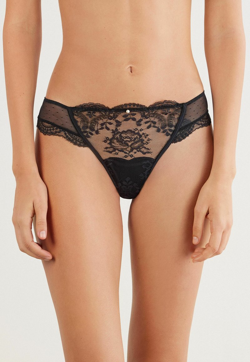 Zwarte kanten strings met doorzichtige gaasdelen, gegolfde randen en bloemmotieven. Inclusief een kleine decoratieve accent aan de voorkant.