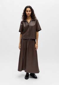 Femme aux cheveux bouclés portant un haut en cuir marron à manches courtes et une jupe longue assortie, associée à des chaussures noires à lacets, sur un fond blanc.