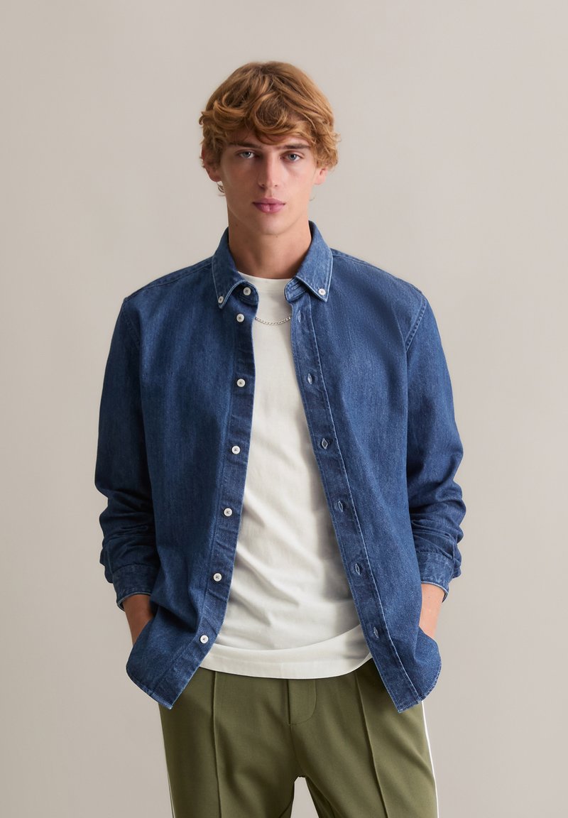 Denim overhemd in donkerblauw, met een klassieke kraag, lange mouwen en witte knopen, gedragen over een effen wit T-shirt.