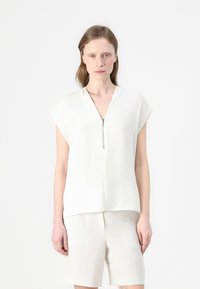 Camisa blanca de manga corta con escote en V y cremallera en la parte frontal. Hecha de una tela suave con un corte holgado, combinada con pantalones cortos de colores claros.