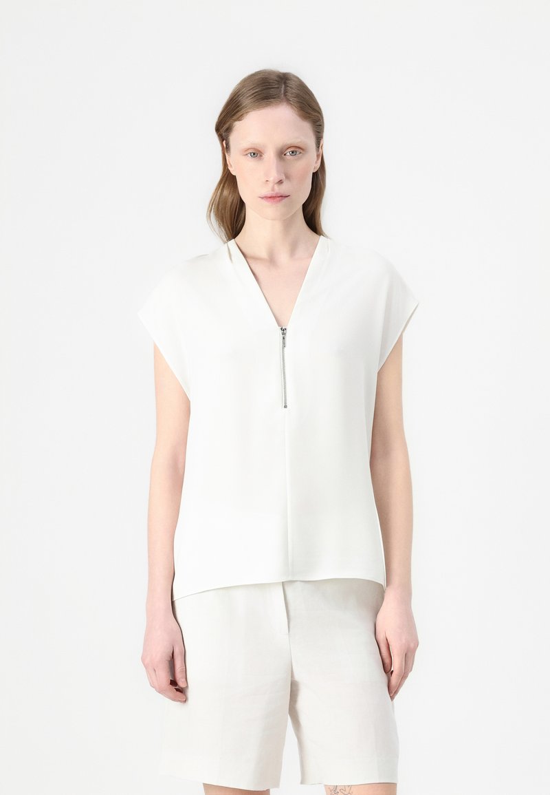 Camisa blanca de manga corta con escote en V y cremallera en la parte frontal. Hecha de una tela suave con un corte holgado, combinada con pantalones cortos de colores claros.