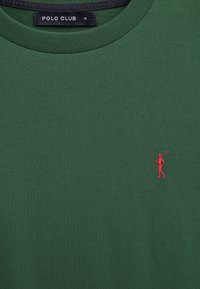 Maglietta verde del Polo Club con scollo rotondo, etichetta di taglia media e piccolo logo ricamato di un giocatore di polo rosso sul petto.