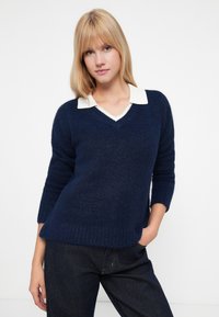 Pulover bleu marin, cu decolteu în V, cu mâneci lungi și tiv striat, suprapus peste o bluză albă cu guler. Material moale texturat.